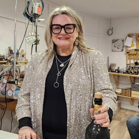 Mrs LollaMac herself, standing behind the shop counter. She is wearing a very sparkly silver jacket and holding a bottle of fizz.