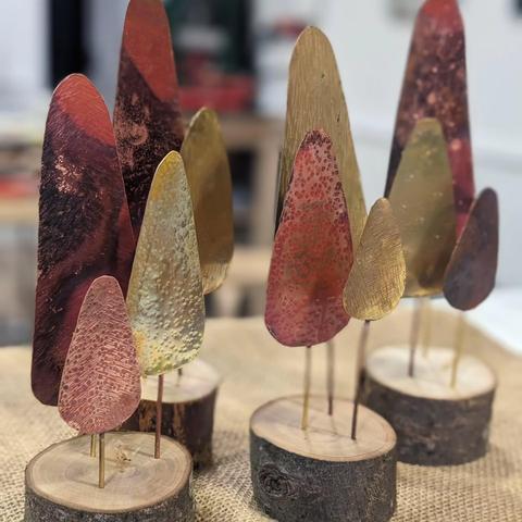 Tree decorations made of a circular slice of wood, with trees formed of copper or brass on metal posts. The metal trees are coloured from being heated for soldering.