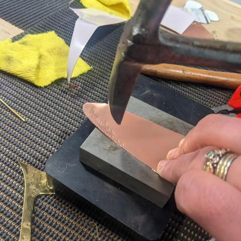 A hand holding a piece of copper on a hammering block, and hitting it with a pointed hammer to give it texture for forming a tree.
