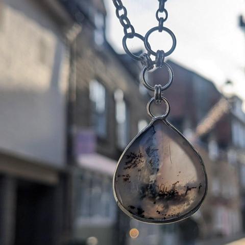 A large white-ish teardrop stone with black inclusions set in a silver bezel with an open back so some light can come through. It is hanging from some silver loops as part of a necklace. In the blurred background are stone buildings.