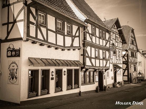 Sepiafarbenes Straßenbild mit einer Reihe historischer Fachwerkhäuser in der Laubacher Altstadt. Im Vordergrund die Gaststätte „Zur Eule“ mit tiefen Fenstern, Blumenkästen und altem Wirtshausschild. Die warme Tönung verleiht der Szene einen nostalgischen, erinnerungsvollen Charakter, der an frühere Zeiten und persönliche Geschichten erinnert.