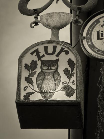 Historisches Wirtshausschild mit einer gemalten Eule, umgeben von Eichenblättern, montiert an einer alten Metallhalterung. Das Schild leuchtet warm vor einem hellen Himmel und vermittelt eine nostalgische, fast märchenhafte Stimmung. Symbol des traditionsreichen Gasthauses „Zur Eule“ in Laubach.