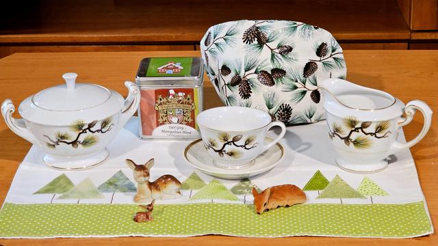 Tea tableau in green and white with images of trees and pinecones on linen and china, and with a trio of vintage deer figurines. The tea is Metropolitan Earl Grey from English Tea Store.