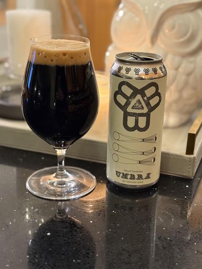 A tall snifter glass full of dark beer with a bubbly tan head. The tan can has a diagram of the moon casting varied shadows in an eclipse