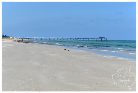 A tranquil colour photograph capturing the wide, sandy beach and the historic Semaphore Pier in Adelaide, South Australia.

The long, horizontal pier extends over the turquoise water of Gulf St Vincent into the distance under a vast, cloudless light blue sky.

The foreground is dominated by the pale, sun-drenched sand, with the water gently lapping the shore.