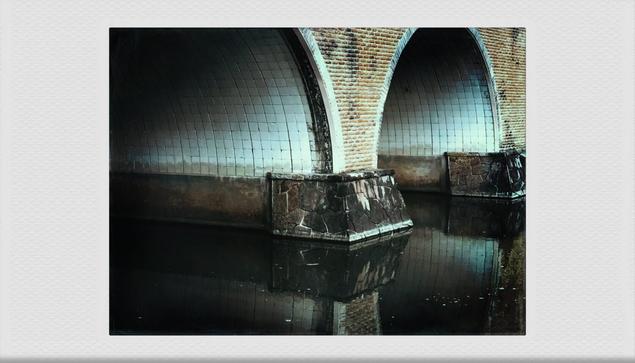 A color photo showing a cropped view of an arched bridge over a canal. The arches of the bridge are tiled and reflected in the dark canal water below.