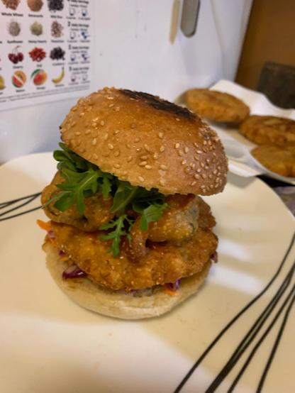 A toasted sesame bun with slaw, chickpea hush puppies, ketchup, tofu phish, vegan tartar sauce, and arugula on a white plate.