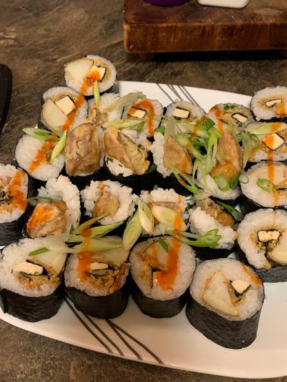 Vegan beer-battered phish and potato wedges wrapped in rice and nori, sliced into rounds and sitting on a white plate, sprinkled with green onions and drizzled with sriracha.