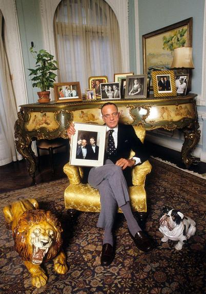 An older man in a suit, holding a picture of himself and Trump. He is sitting in a golden chair, surrounded by golden furniture and a fake lion (and real dog?).