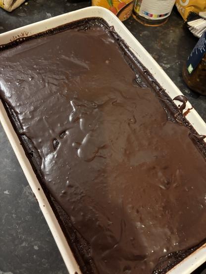 A large rectangular tray of chocolate cake covered in a smooth, glossy layer of melted chocolate topping. The surface is shiny with visible swirls and streaks from spreading, and the dark chocolate glaze reaches right to the edges of the pan. The cake is set on a dark kitchen countertop with jars and containers visible in the background.