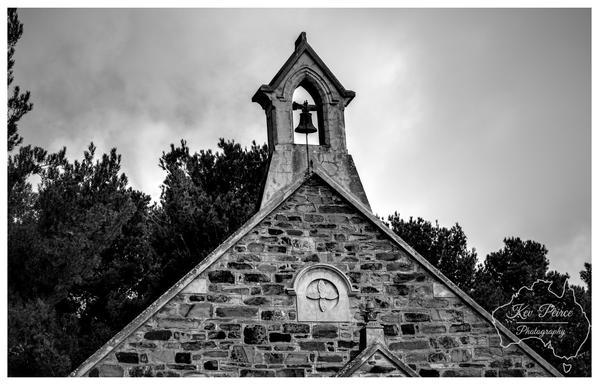 A dramatic black and white photograph, captured at a low angle, focusing on the stone gable and bell tower of an historic church in Burra, South Australia.  The wall is constructed of rough hewn stone blocks. A circular carving is visible in the center of the gable.  The bell tower arch contains a small, dark bell. Dense, dark foliage from surrounding trees frames the structure against a bright, cloudy sky.