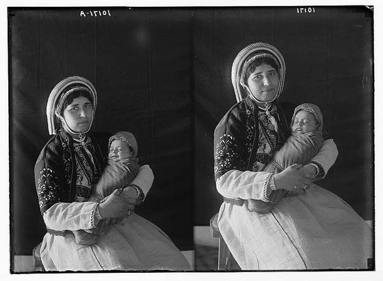 This black and white image depicts two photographs placed side by side. Both show a woman dressed in traditional attire, with embroidered details on her jacket and headscarf. She is holding an infant wrapped in cloth, both of them appearing to be engaged in quiet contemplation or restful observation.
The left photograph captures the moment from slightly higher angle where she looks directly at the camera while cradling the child close. In contrast, the right image shows a lower-angle shot with her gaze directed downwards towards the sleeping infant on her lap. The background is nondescript and dark, ensuring that focus remains on the subjects.
The top of each photograph has handwritten markings "A-110[illegible]", suggesting it might be part of a series or collection. Additional context indicates this image could have historical significance as it seems to depict traditional attire possibly associated with Palestinian culture during the early 20th century, and is linked to American Colony in Jerusalem.
The photo credits belong to Eric and Edith Matson who were photographers known for their documentation of Palestine at that time period. The provided link suggests this image might be part of a larger collection held by "loener.nl," which could imply it's available through an online archive or database dedicated to the works of these photographers.
It is important to note that any attempt to speculate beyond what can clear [...]