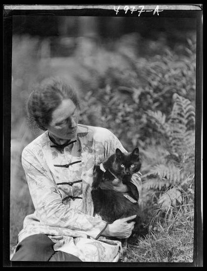 The image is a black and white photograph featuring an older woman seated outdoors, holding a cat in her arms. The woman appears to be wearing glasses and has short hair with some strands of gray visible on the sides. She's dressed in what looks like a Chinese-style jacket adorned with floral patterns.

She holds a dark-colored domestic cat close to her chest; the feline gazes slightly upwards at the camera, giving an impression that it is quite relaxed or perhaps curious about its surroundings and possibly interested in being photographed.

The background of the photograph shows foliage typical of temperate climates - ferns and other green plants suggest this may have been taken during a warmer season. The photo seems to be well-composed with attention given to framing, as there's enough detail around her without distracting from her or the cat she is holding.

There are handwritten annotations in white ink on top: "4972-A," which could indicate cataloging information for this particular photograph within an archive. Below that text, it reads "Miss" and then seems incomplete with a dash followed by another word starting with 'f', possibly referring to the woman's name or her status.

The image has been referenced in various records as belonging to Arnold Genthe (1869-1942), known for his work capturing historical moments including events such as World War II, but it is also linked to a different individual named Gabriel [...]