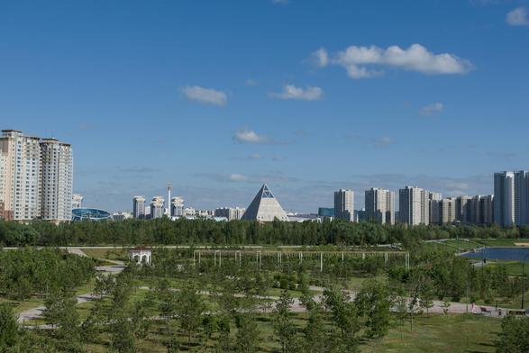The image captures a panoramic view of Astana, Kazakhstan, with the Palace of Peace and Reconciliation in the center. The focal point is an imposing pyramid-like structure with a triangular facade and white exterior, standing majestically amidst lush greenery. In contrast to its geometric simplicity, the skyline behind it boasts modern architecture, including skyscrapers reaching towards blue skies dotted with fluffy clouds.

The colors in the image are vibrant yet harmonious, dominated by shades of green from the surrounding trees and grassy areas, white and gray tones reflecting off the buildings' facades, and a clear blue sky. The perspective is such that it provides an expansive view of both the foreground - featuring man-made structures amidst nature - and the background - showcasing urban development under the open skies.

This image codeifies not just physical objects but also human engineering marvels in harmony with natural beauty.