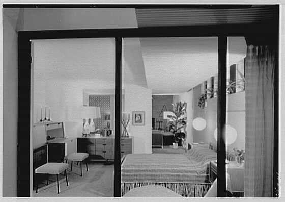 The image is a black and white photograph depicting an interior living space, likely from the mid-20th century. It shows a large room with modern furnishings including upholstered sofas, chairs, a coffee table, lamps, and various decorative items such as vases, candles, paintings, and plants. The layout suggests an open-plan design characteristic of post-war suburban homes in America.
The perspective is from within the living space looking out through glass doors or windows that separate it from another area with visible artwork on the wall. Light streams into the room, reflecting off shiny surfaces like chrome vases and enhancing the overall brightness. Decorative patterns are noticeable on some textiles such as a sofa throw and possibly curtains.
This image is identified by its reference to "Ludwig Baumann," who appears to be associated with home furnishings exhibits in September 1952. It's part of an exhibit managed by Gottsch- Schleisner, Inc., suggesting it was taken at or near such a display event during that time period.
The photograph carries historical significance as it captures the style and ambiance typical for American living spaces from this era, likely intended to showcase modern interior design trends or products available in home furnishing markets.