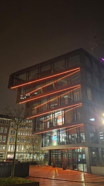 Foto eines Bürogebäudes. An der gestuften Glasfassade sind über die gesamte Breite des Gebäudes horizontale Lichtbänder installiert, die orange leuchten. Die Streifen spiegeln sich in der Fassade. Auch der Gehweg vor dem Gebäude reflektiert das orange Licht.