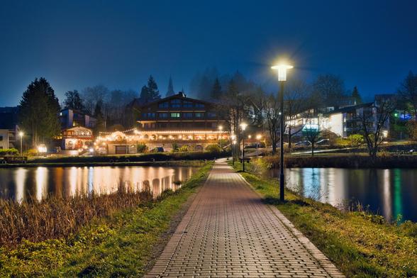 1. Der Vordergrund und die Perspektive
Der Weg: Zentral im Bild führt ein gepflasterter Weg aus rötlichen Steinen geradeaus in die Tiefe. Er dient als perfekte Führungslinie, die den Blick des Betrachters direkt zum Hauptgebäude im Hintergrund lenkt.
Vegetation: Links und rechts des Weges wächst Gras und trockenes Schilf, das im künstlichen Licht golden und braun schimmert.
2. Die Beleuchtung und das Wasser
Die Laterne: Auf der rechten Seite dominiert eine helle Straßenlaterne den Vordergrund. Durch die vermutlich lange Belichtungszeit entsteht um das Licht herum ein deutlicher „Sternchen-Effekt“ (Blendenstern).
Das Gewässer: Der Weg führt über oder entlang eines ruhigen Gewässers (ein Teich oder kleiner See). Die Wasseroberfläche wirkt durch die Langzeitbelichtung sehr glatt und seidig.
Reflektionen: Die Lichter der Gebäude und der Laternen spiegeln sich langgezogen und malerisch im dunklen Wasser.
3. Der Hintergrund und die Architektur
Das Hauptgebäude: Im Zentrum steht ein großes, mehrstöckiges Gebäude, das wie ein Hotel oder ein gut besuchtes Gasthaus wirkt. Es hat eine breite Fensterfront und eine Terrasse.
Festliche Beleuchtung: Eine Lichterkette zieht sich über die Terrasse des Gebäudes und sorgt für eine warme, einladende Atmosphäre („Gemütlichkeit“).
Umgebung: Man sieht weitere Gebäude links und rechts, die ebenfalls beleuchtet sind. Im Hintergrund ragen Nadelbäume in den Himmel, und es scheint leichter Nebel oder Dunst zwischen den Bäumen und Hügeln zu liegen, was d