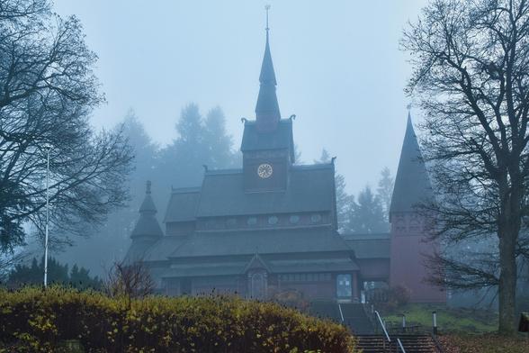 Dieses Bild zeigt eine völlig andere, aber ebenso faszinierende Stimmung wie das vorherige. Wir sehen eine imposante Holzkirche, die in dichten Nebel gehüllt ist. Die Szene wirkt mystisch, fast wie aus einem Märchen oder einem Fantasy-Film.
Hier ist eine detaillierte Beschreibung der Szene:
1. Die Architektur (Das Hauptmotiv)
Baustil: Es handelt sich um eine beeindruckende Holzkirche im Stil einer nordischen Stabkirche. Charakteristisch sind die gestaffelten, schindelgedeckten Dächer, die sich kaskadenartig nach oben verjüngen.
Der Turm: In der Mitte ragt ein hoher, spitzer Turm auf. Darunter befindet sich eine Turmuhr mit goldenem Ziffernblatt, die im trüben Licht fast zu leuchten scheint.
Details: An den Dachfirsten sind verzierte Endstücke zu erkennen (möglicherweise Drachenköpfe oder ähnliche Ornamente), was typisch für diesen Baustil ist. Rechts neben dem Hauptgebäude steht ein weiterer, schlanker Turm.
Material: Das gesamte Gebäude scheint aus dunklem Holz gefertigt zu sein, teilweise rötlich-braun gestrichen, was einen schönen Kontrast zum grauen Nebel bildet.
2. Wetter und Atmosphäre
Der Nebel: Dichter, bläulich-grauer Nebel umschließt das Gebäude und verschluckt den Hintergrund fast vollständig. Dies isoliert die Kirche optisch und lässt sie noch monumentaler erscheinen.
Stimmung: Das Bild strahlt eine gewisse Melancholie und Stille aus. Es wirkt „nordisch“, kühl und geheimnisvoll.
3. Vordergrund und Umgebung
Rahmung: Zwei große, laublose Bäume links und rechts rahme
