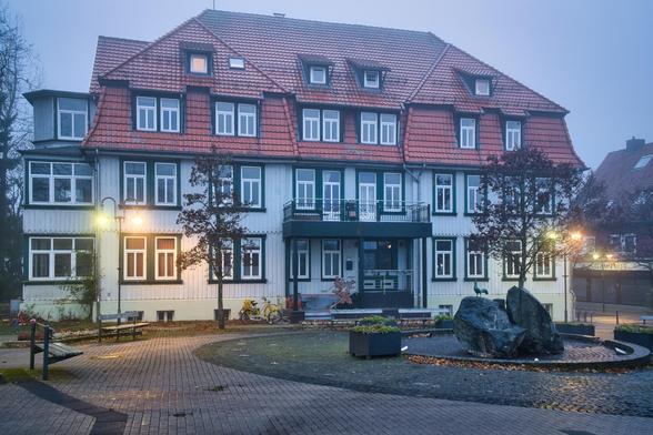 1. Das Hauptgebäude (Architektur)
Imposanter Bau: Das Bild wird von einem großen, breiten Gebäude dominiert, das typisch für die Kur-Architektur im Harz ist. Es wirkt wie ein Hotel, ein Kurhaus oder ein Verwaltungsgebäude.
Fassade: Die Fassade ist weiß verkleidet (wahrscheinlich Holzlatten), was durch dunkelgrüne Fensterrahmen und Balken akzentuiert wird.
Dach: Ein mächtiges, rotes Ziegeldach mit vielen Dachgauben deckt das Haus.
Symmetrie: Der Bau ist weitgehend symmetrisch angelegt, mit einem zentralen Eingangsbereich, der durch einen kleinen Vorbau und einen darüberliegenden Balkon betont wird.
2. Der Vorplatz und Details
Pflasterung: Im Vordergrund sieht man einen gepflasterten Platz. Die Steine glänzen feucht, was darauf hindeutet, dass es geregnet hat oder der Nebel sehr nass ist.
Der Brunnen: Rechts im Vordergrund steht ein markanter Brunnen, der aus großen, groben Felsbrocken besteht. Darauf thront eine kleine Statue – wenn man genau hinsieht, erkennt man einen Hahn. (Ein schönes Detail, da der Ort höchstwahrscheinlich Hahnenklee ist).
Das gelbe Fahrrad: Links neben dem Eingang lehnt ein leuchtend gelbes Fahrrad (evtl. ein Postrad oder Zustellrad). Es ist der einzige kräftige Farbtupfer im sonst eher gedeckten Bild.
Möblierung: Mehrere schlichte Holzbänke stehen auf dem Platz verteilt und wirken in der nebligen Szenerie etwas verlassen.
3. Licht und Atmosphäre
Wetter: Es herrscht dichter Nebel. Der Hintergrund, inklusive der Bäume links und der Gebäude rechts, verblas