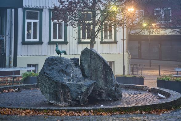 Das Bild zeigt eine Detailansicht der Szene, die wir gerade im Weitwinkel betrachtet haben. Hier liegt der Fokus ganz klar auf dem Brunnen im Vordergrund.
Hier ist die genaue Beschreibung:
1. Das zentrale Motiv: Der Hahnen-Brunnen
Die Felsen: In der Mitte steht eine Formation aus massiven, groben Felsbrocken (vermutlich Granit oder ein ähnliches Gestein). Sie wirken schwer und dunkel, was durch die Nässe noch verstärkt wird.
Der Hahn: Auf dem linken, etwas flacheren Felsen thront eine grünliche Metallfigur (wahrscheinlich oxidiertes Kupfer oder Bronze), die einen stolzen Hahn darstellt. Dies ist ein direktes visuelles Zitat des Ortsnamens (Hahnenklee).
Beleuchtung: Am Boden um die Felsen herum sind kleine Strahler installiert, die darauf hindeuten, dass der Brunnen nachts gezielt angestrahlt wird.
2. Vordergrund und Boden
Herbststimmung: Der Boden ist mit braunem und gelbem Herbstlaub bedeckt, das sich am Rand des gepflasterten Kreises sammelt.
Nässe: Das Kopfsteinpflaster glänzt stark, was die feuchte, nasskalte Witterung unterstreicht.
3. Der Hintergrund
Architektur:
Links: Man sieht detailliert die Fassade des weißen Holzhauses mit den dunkelgrünen Fensterrahmen.
Rechts: Im Hintergrund verschwimmt ein weiteres Gebäude im Nebel. Man erkennt den beleuchteten Schriftzug eines Cafés oder Restaurants ("AVANTI ... TREFF"), dessen Schaufenster warmes Licht ausstrahlen.
Vegetation: Ein Baum mit spärlichem, rötlichem Restlaub steht direkt hinter dem Felsen und ragt in den oberen Bi