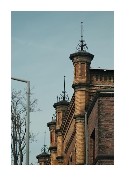 Detailansicht eines historischen Backsteingebäudes mit mehreren turmartigen Aufsätzen. Auf den kleinen Türmchen sitzen filigrane metallene Spitzen und ornamentale Verzierungen. Rechts ist die Fassade aus rotbraunem Backstein zu sehen, links ragen kahle Äste eines Baums und ein Teil eines Metallpfostens ins Bild. Der Himmel ist blassblau und wolkenlos.