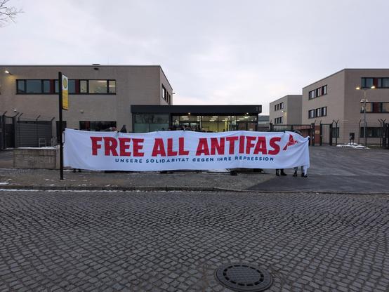 Banner mit der Aufschrift 'Free all antifas' vor bunkerartigen Gebäuden. Strasse vor dem Banner ist gepflastert.