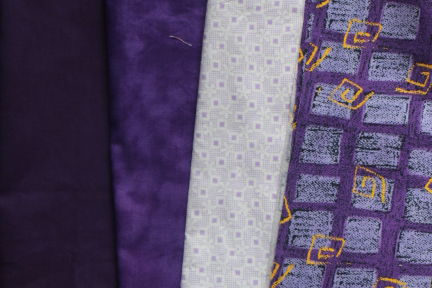 Assorted purple fabrics group together. One of the fabrics has yellow square swirls as a pattern over the light purple rectangles.