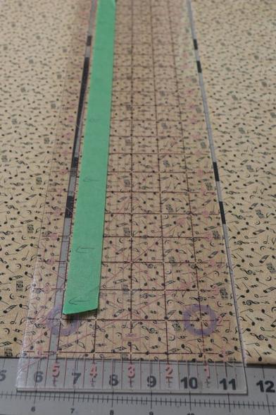 Cutting ruler on top of some beige fabric. Masking tape is stuck to the ruler at the 5 mark. There are arrows drawn on the masking tape pointing towards the 5 mark. This is so I dont cut the fabric strips too thin. I need them chunky for this project.