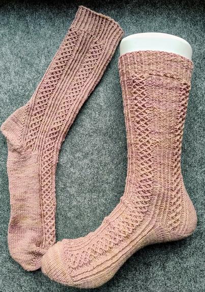 A pair of handknit socks in pale pink.

One sock is on a plastic mannequin, the other is laid out next to it on a grey background

The socks feature columns of lattice broken up by vertical lines knit stitches.