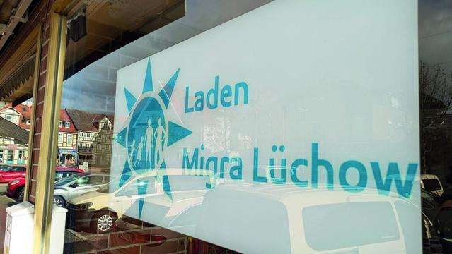 Glasfront eines Geschäfts mit Aufschrift "Laden" und dem Schriftzug "Migra Lüchow", Spiegelung von Fahrzeugen und Gebäuden im Glas.