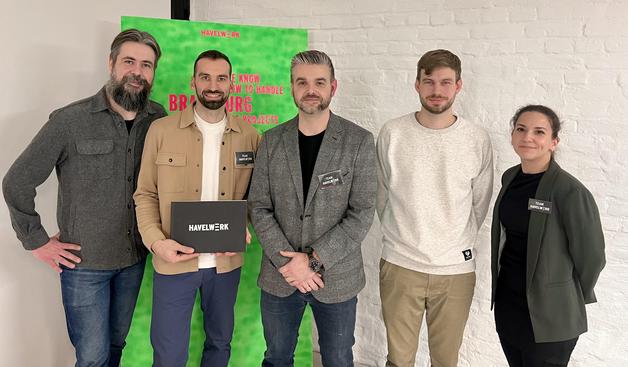 Eine Gruppe von fünf Personen steht vor einem bunten Hintergrund mit dem Schriftzug „HAVELWERK”. Sie posieren gemeinsam, wobei eine Person eine schwarze Box mit dem gleichen Namen in der Hand hält.