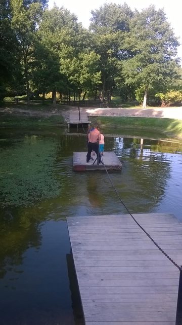 Photo from a parks pond, with a chain between two small docks with stairs, where you can pull yourself on a small platform across it. The platform is in the middle, with a shirtless man and some small kids pulling themselves across the small pond.