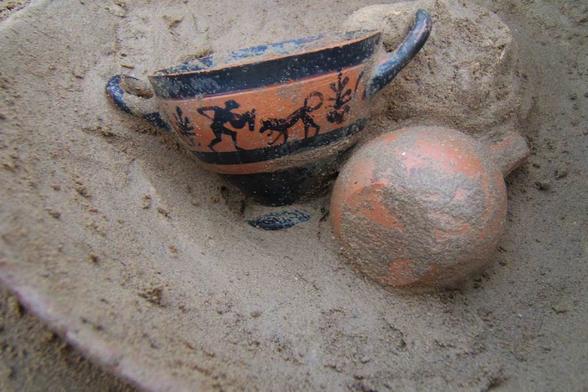 Due reperti archeologici, tra cui una coppa decorata con figure umane e un vaso, parzialmente sepolti nella sabbia.