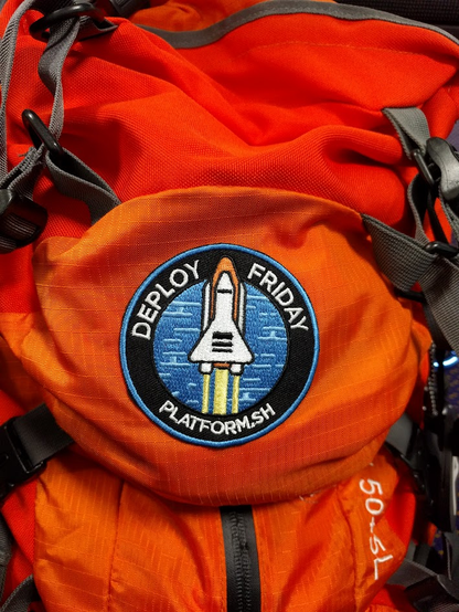 Orange back with a round patch like sticker that says deploy Friday, with a space shuttle still with booster rockets and main orange tank attached mid launch.