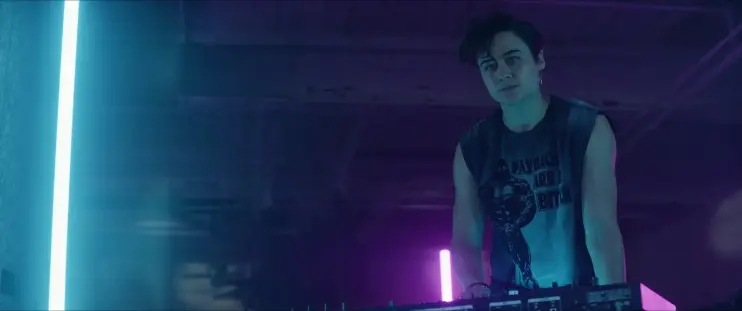 Image de Queens of the dead. Dre aux platines de son club, en contre-plongée. L'éclairage vient de barres néons verticales, on en voit une bleue et une violette.