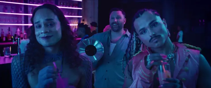 Image de Queens of the dead. Dans le club (éclairage néons violet et bleu), Nico au premier plan avec un verre à cocktail aux cotés de son ami.e Twiz qui sourit, son propre cocktail à la main. Derrière, Barry, nettement très cis et hétéro par rapport à tout le reste des personnages, une poubelle sous le bras, a l'air de bien se marrer.