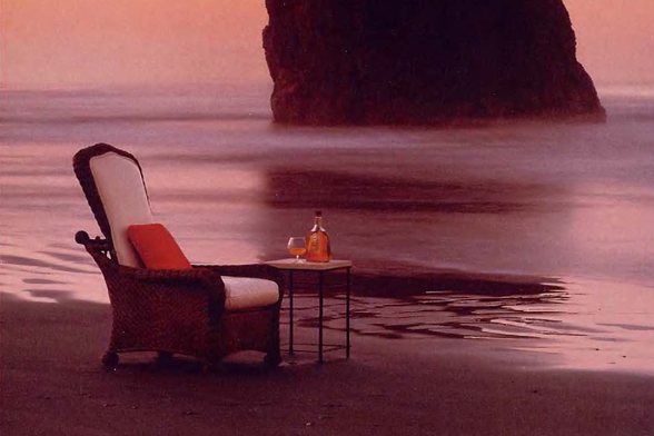 1992 A Serene Evening by the Sea

The image captures a tranquil moment at dusk, as soft pink and orange hues paint the sky above.
On a deserted beach, an armchair with plush red cushions sits on sandy terrain adjacent to calm waters where a large rock formation emerges from the sea.