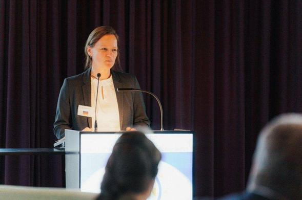 Eine Frau hält eine Rede an einem Rednerpult. Sie trägt einen dunklen Blazer über einer hellen Bluse. Auf ihrer linken Seite ist ein Namensschild befestigt. Das Rednerpult ist weiß und hat eine schwarze Oberfläche, auf der zwei Mikrofone stehen. Dahinter hängt ein dunkelroter Vorhang. Im Vordergrund sind unscharf die Köpfe von Zuhörern zu sehen. Die Frau scheint in einem professionellen Umfeld zu sprechen, möglicherweise auf einer Konferenz oder einer ähnlichen Veranstaltung.