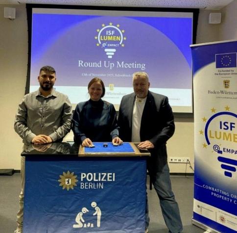 Drei Personen posieren für ein Foto vor einer Leinwand und einem Tisch mit der Aufschrift "Polizei Berlin". Die Leinwand zeigt das "ISF Lumen Empact Round Up Meeting" vom 13. November 2025 in Schwäbisch. Links steht ein Mann mit Bart, in der Mitte eine Frau und rechts ein Mann im Sakko. Auf dem Tisch liegen kleine Gegenstände. Ein Banner rechts wirbt ebenfalls für "ISF Lumen Empact" und "Combatting Organized Property Crime". Das Projekt wird von der Europäischen Union und Baden-Württemberg kofinanziert.