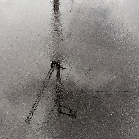 Square format. Wet tarmac and a small puddle catch the reflection of a telegraph pole and attendant wires. The pole enters the frame from the top edge and extends through the centre. Wires project at 3, 7 and 10 o'clock but their clarity breaks up as they pass through slightly less saturated areas. Although a color shot, the scene is monotone due to overcast skies above. A single raindrop has hit the puddle and small ripples echo.