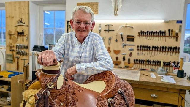 Mann in Hemd vor Werkbank mit aufwendig verziertem Sattel, Werkzeuge an der Wand, heller Raum.