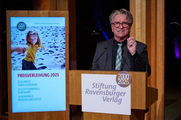 Minister Manne Lucha bedankt sich bei der Stiftung Ravensburger Verlag für 25 Jahre Engagement für Kinder und Jugendliche. Er steht dabei hinter einem Rednerpult auf der Bühne.