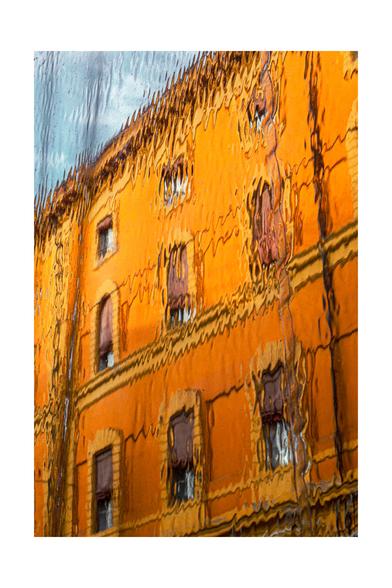 Photograph of an orange-painted building through a plastic rain cover which gives the appearance of peeling paint and visible cracks. The plastic sheet distorts the building with ripples and waves. The building has three rows of windows.The sky in the background is a soft blue, contrasting with the warm, worn tones of the building.