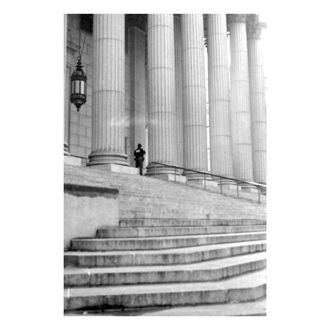 A police officer stands atop the steps of the New York County Supreme Court. Standing there, being shrunken by the large pillars of the building.