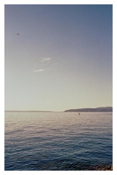 Color photo of the Adriatic Sea with a view of the island of Cres, taken from a swimming spot between Ika and Icici. Although it was taken in the morning, the light is already very bright. The sky takes up almost two-thirds of the image, ranging from blue to almost white on the horizon. A small boat floats in the water. A bird flies into the picture from the left.
