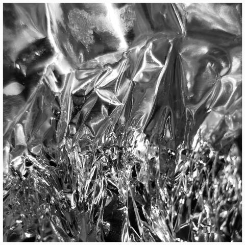 Black-and-white capture of a shiny silvery metallic foil that is crumpled. White light reflections accentuate the sharp, zigzag structure of the material, which looks like broken ice floes in moving water. Some ice floes pile up on top of each other and are pushed upwards, while another slow, icy wall of water approaches from the background. It will bury and drown everything beneath it.