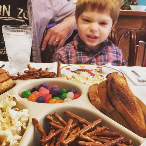 Color image of serving a Thanksgiving meal to a toddler in the manner of the cartoon classic, A Charlie Brown Thanksgiving, complete with toast, pretzels, popcorn and jelly beans as the entire meal, just like the Peanuts kids get in the cartoon. The toddler was both confused and not amused.  Image shows a toddler looking at a table server full of popcorn, pretzel, toast and jelly bean selections only in place of the traditional American Thanksgiving dinner of turkey, stuffing, gravy, mashed potatoes, sweet yams, green beans, cranberry sauce, and more.