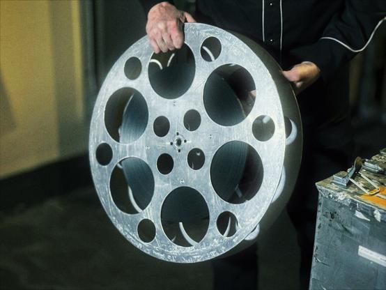 Large film canisters, each labeled a number from "1" to "10," sit on a small cart in a film storage room of the Hollywood Theatre, with one of the reels being held for the camera.