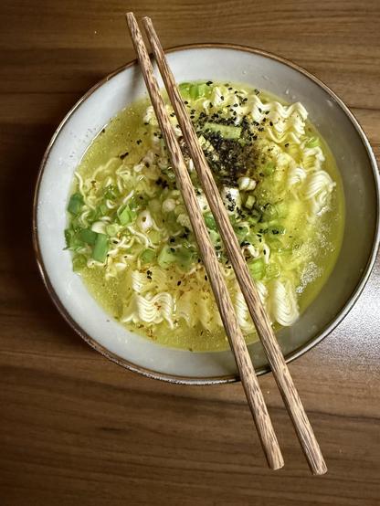 Eine Schüssel Ramen-Nudeln in einer leichten Brühe, garniert mit gehackten Frühlingszwiebeln und mit schwarzem Sesam bestreut. Zwei Holzstäbchen ruhen auf dem Rand der Schüssel. Die Schüssel wird auf eine Holzoberfläche gestellt.
