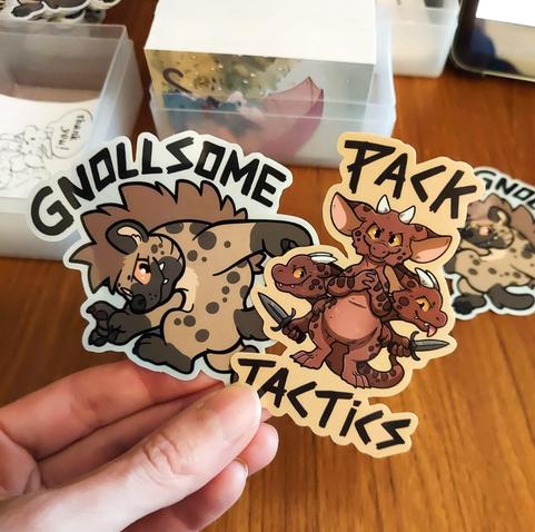 A photo of a hand holding up a gnoll and kobold set of stickers